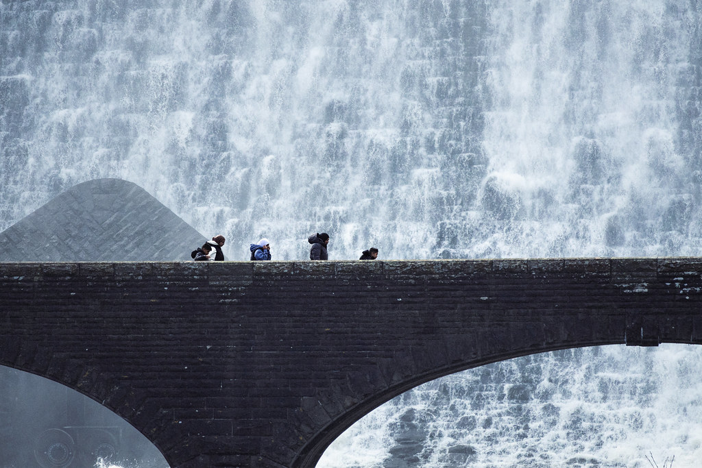 People crossing a stone bridge below a reservoir dam waterfall in Wales